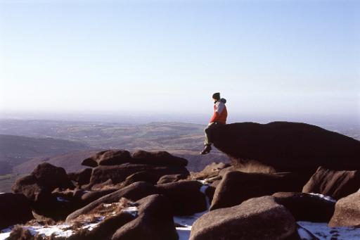 Lone figure of a man sitting on a mountain peak on the edge of a rock with scattered snow overlooking mountain ranges and valleys as he contemplates nature