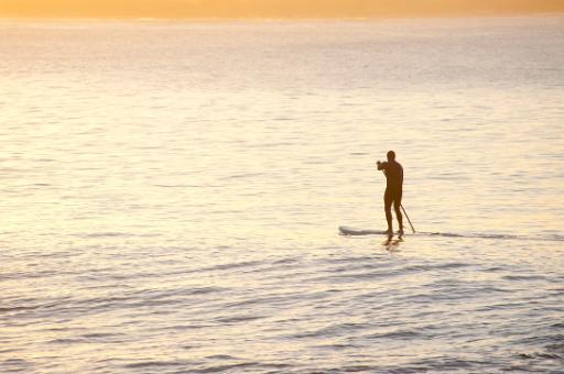 Man on a paddle board paddling across a calm ocean parallel to a sandy tropical beach, silhouetted against bright water with copyspace